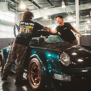 Two men shaking hands next to a black and teal sports car in a showroom.