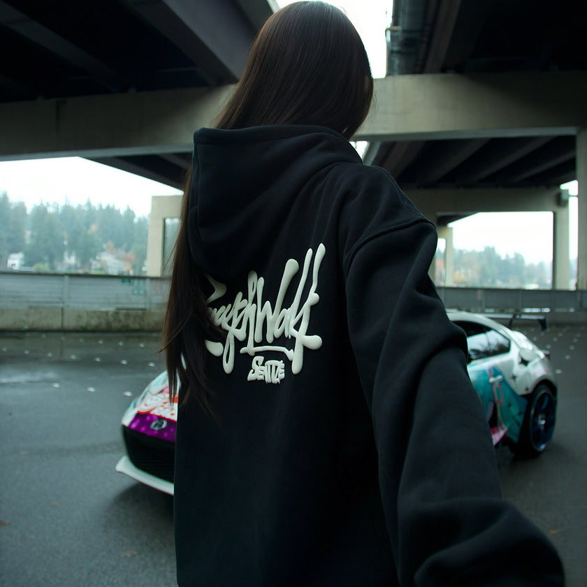 Person wearing a black hoodie with a logo on the back, standing under a bridge with cars in the background.