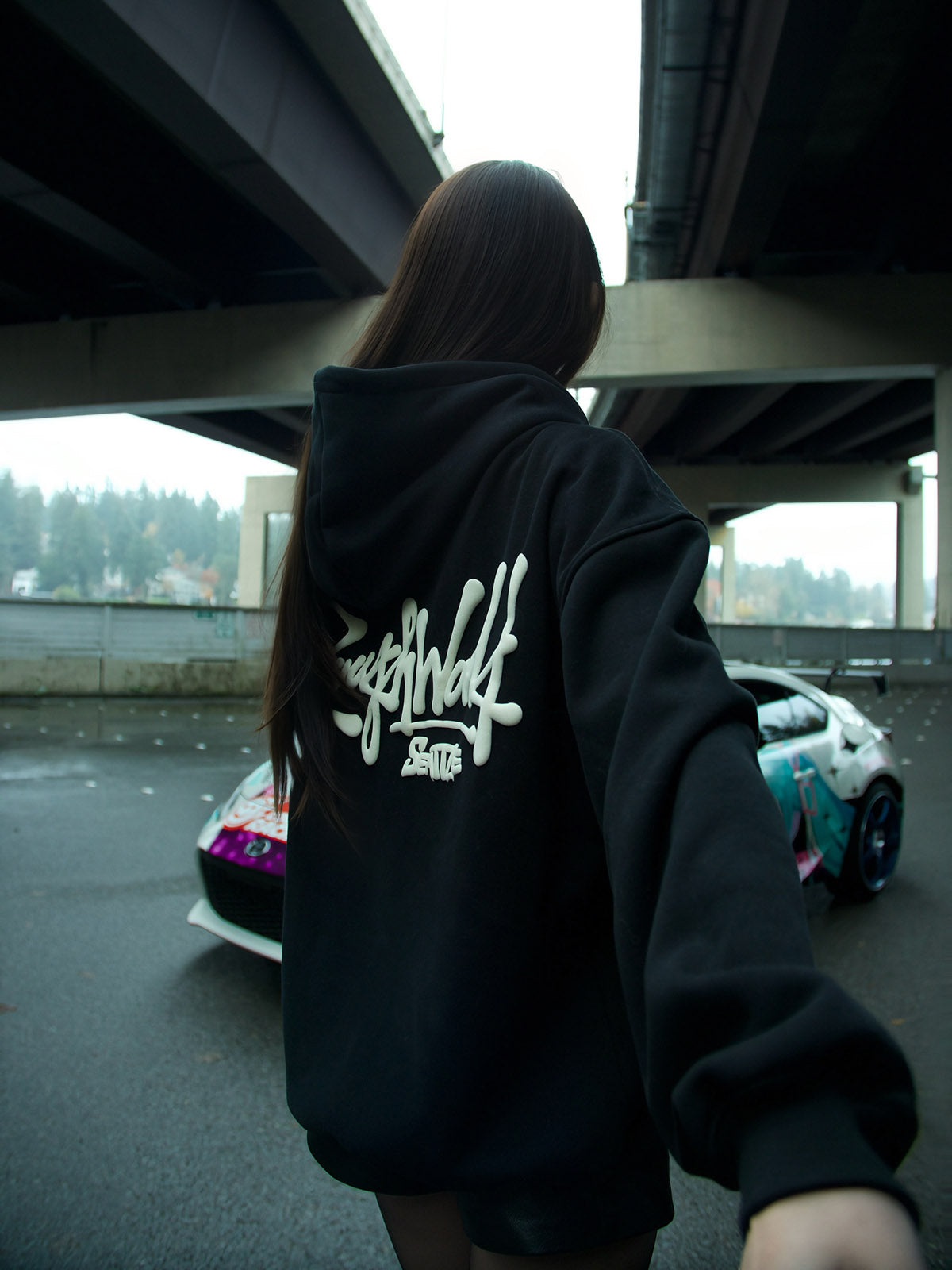 Person wearing a black hoodie with a logo on the back, standing under a bridge with cars in the background.