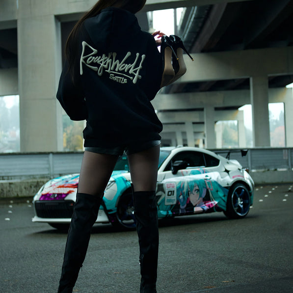 Person wearing a hoodie with 'Rough World' branding, standing in front of a car with anime character design.