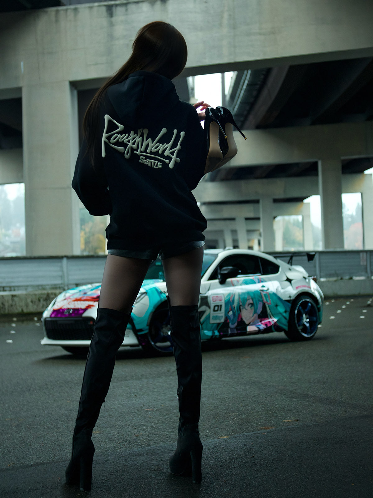 Person wearing a hoodie with 'Rough World' branding, standing in front of a car with anime character design.