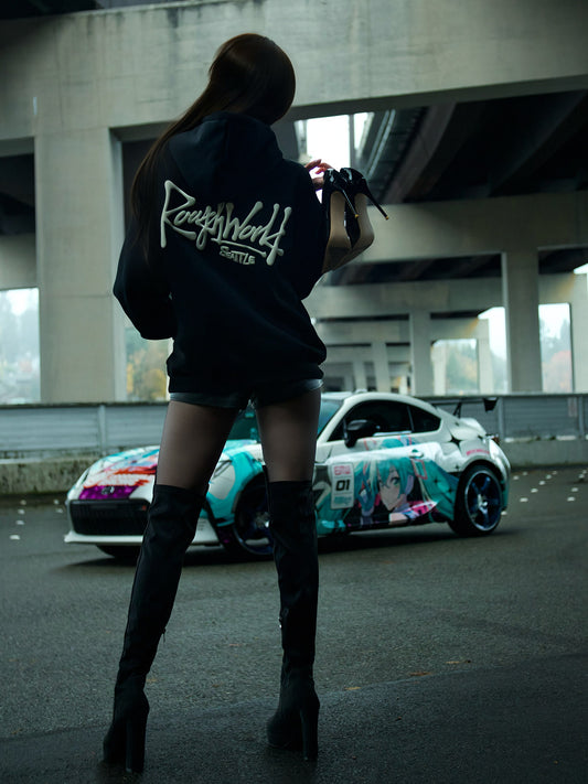 Person wearing a hoodie with 'Rough World' branding, standing in front of a car with anime character design.