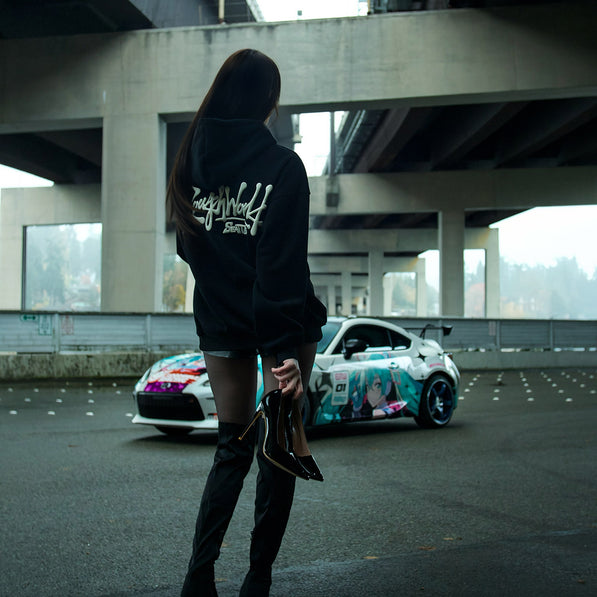 Person wearing a black hoodie with text, standing in front of a colorful car under an overpass.