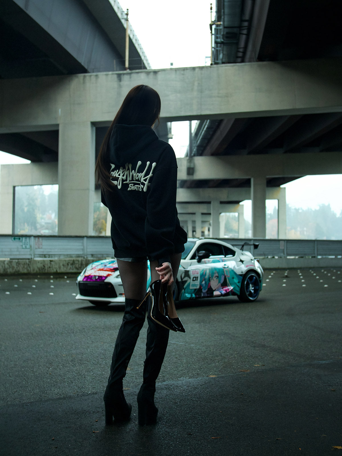 Person wearing a black hoodie with text, standing in front of a colorful car under an overpass.